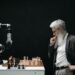 A thoughtful, bearded man in glasses considering a robotic arm by a chess board — the deliberate decision-making UK independent optician practice owners face on AI.