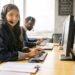Smiling support agent wearing a headset at a computer with a colleague behind — support, onboarding and training in UK optician practice management software.